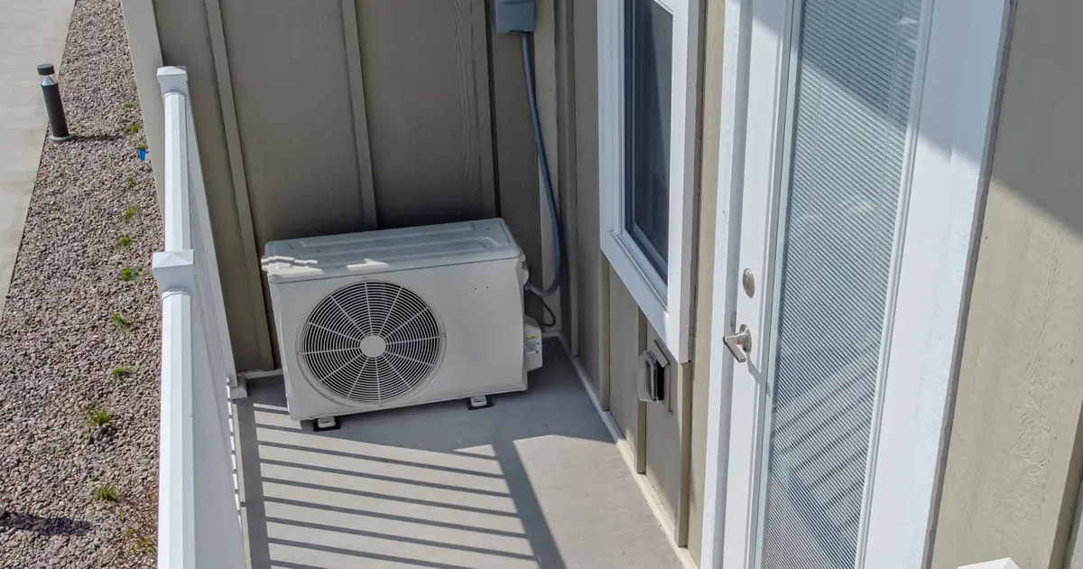 Buitenunit van een warmtepomp zonder merkopdruk op het balkon van een woning, naast de achterdeur